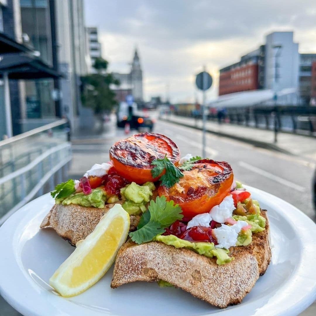 Moose Coffee Avocado Toast Grilled Tomatoes City Background Fresh avocado toast with grilled tomatoes and a lemon wedge served on a white plate at Moose Coffee, with a city street backdrop.