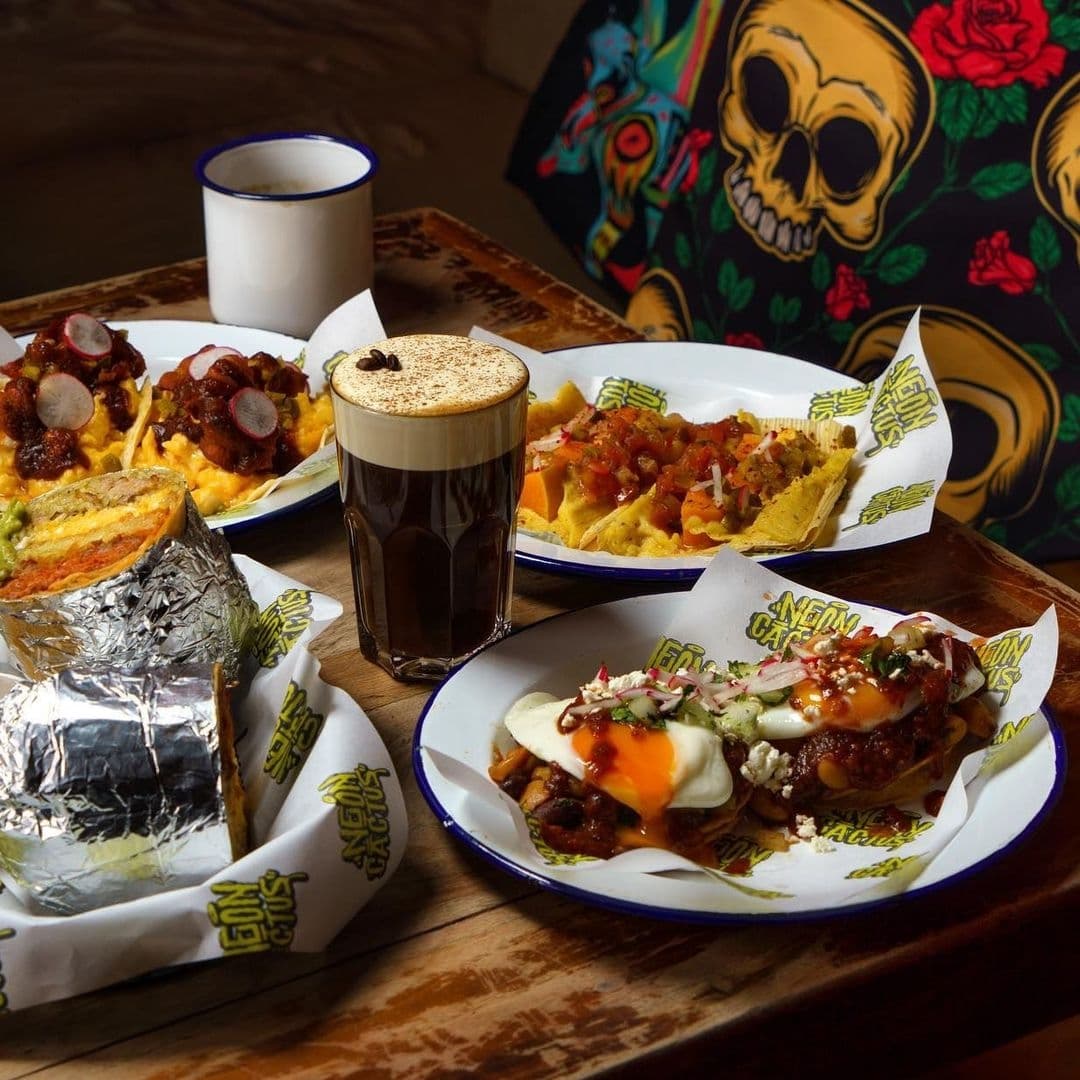 Neon Cactus Mexican Cuisine Colorful Plates Craft Beer Assorted Mexican dishes and a pint of dark beer on a wooden table at Neon Cactus. Visible are nachos with toppings, a burrito wrapped in foil, and huevos rancheros with a sunny-side-up egg. A colorful skull and floral pattern decorates the background.