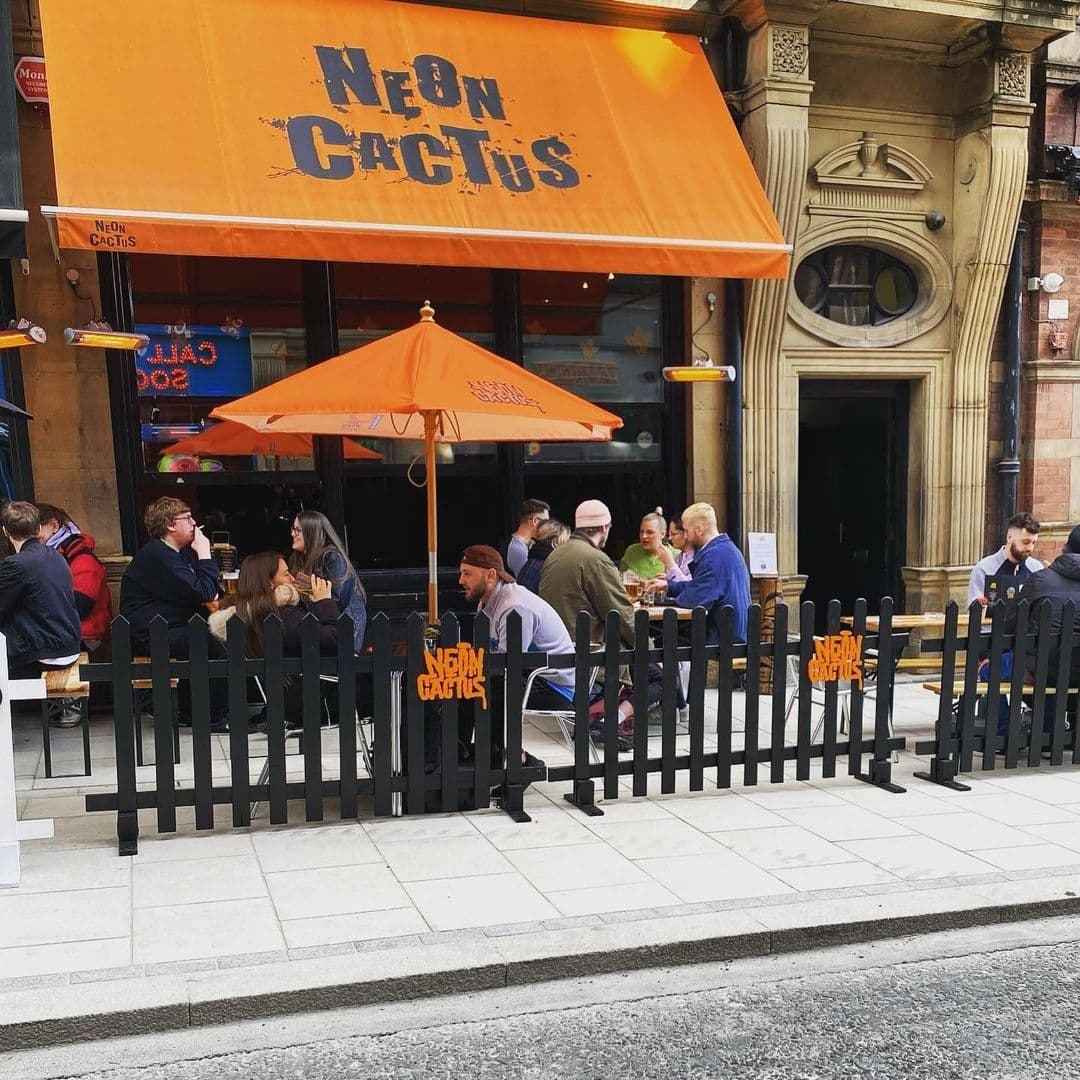 Neon Cactus_Outdoor Dining_Patio Umbrellas_Casual Atmosphere Outdoor seating area at Neon Cactus with patrons dining under an orange umbrella and the restaurant's name displayed on a black fence and awning.