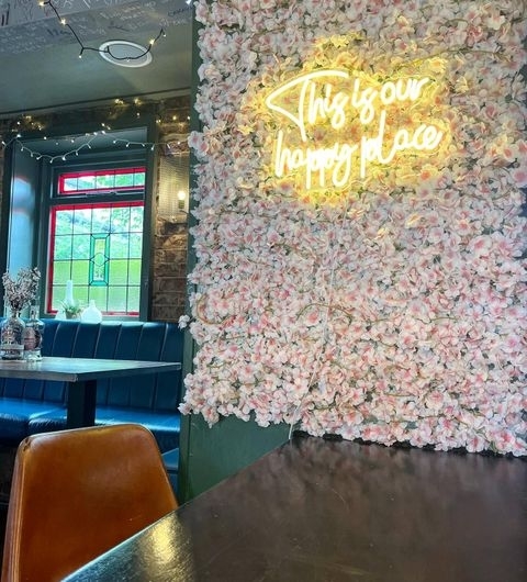 Number Thirty Three_Happy Place Neon Sign Floral Wall Interior of Number Thirty Three featuring a cozy corner with blue seating, a wall adorned with pink flowers, and a neon sign that reads "This is our happy place".
