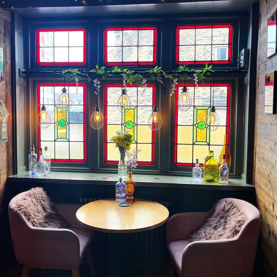 Number_Thirty_Three_Stained_Glass_Windows_Cozy_Interior Cozy interior of Number Thirty Three with a round wooden table, two plush chairs with grey cushions, and a window sill adorned with colorful glass bottles and fresh flowers. The room is lit by stylish hanging bulbs and features large windows with vibrant stained glass panels in red, yellow, and green.