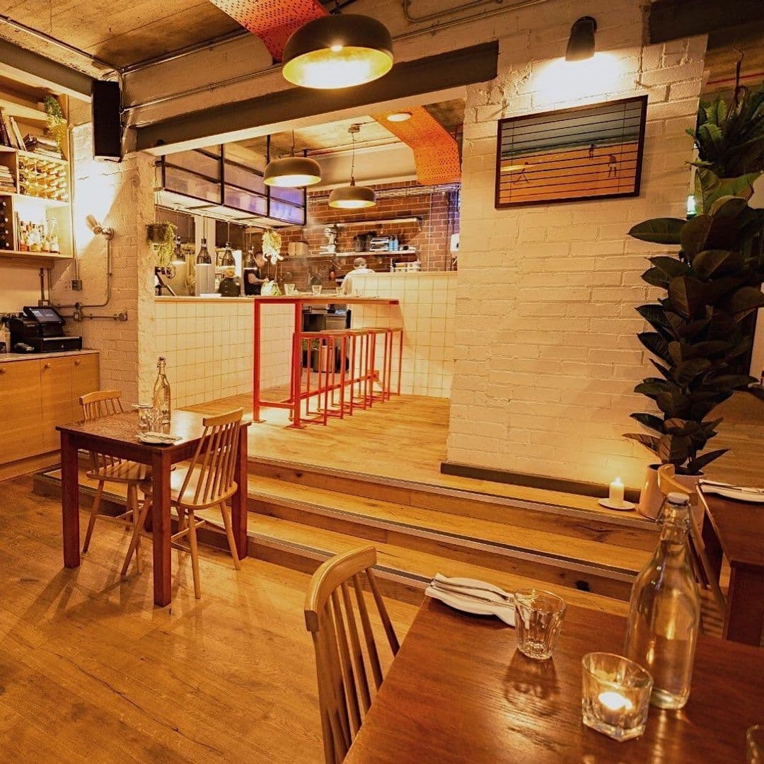 Ox Club at Headrow House Cozy Industrial Dining Interior Interior of Ox Club at Headrow House featuring warm lighting, wooden dining tables, exposed brick walls, and an open kitchen with chefs at work.