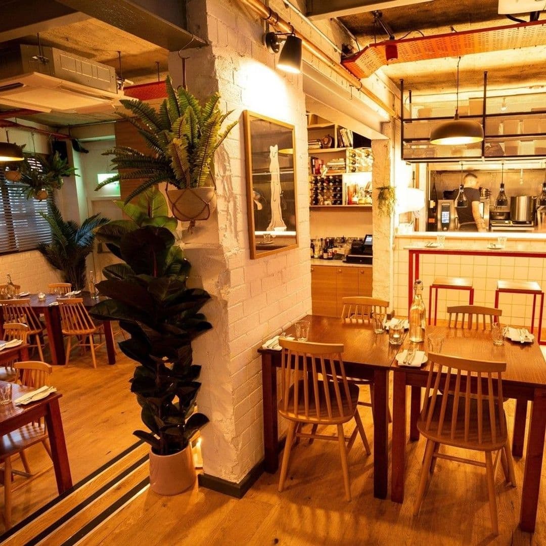 Ox Club at Headrow House Cozy Industrial Dining Space Interior of Ox Club at Headrow House featuring a cozy dining area with wooden tables and chairs, white painted brick walls adorned with artwork, lush green plants, and a well-lit bar area in the background.