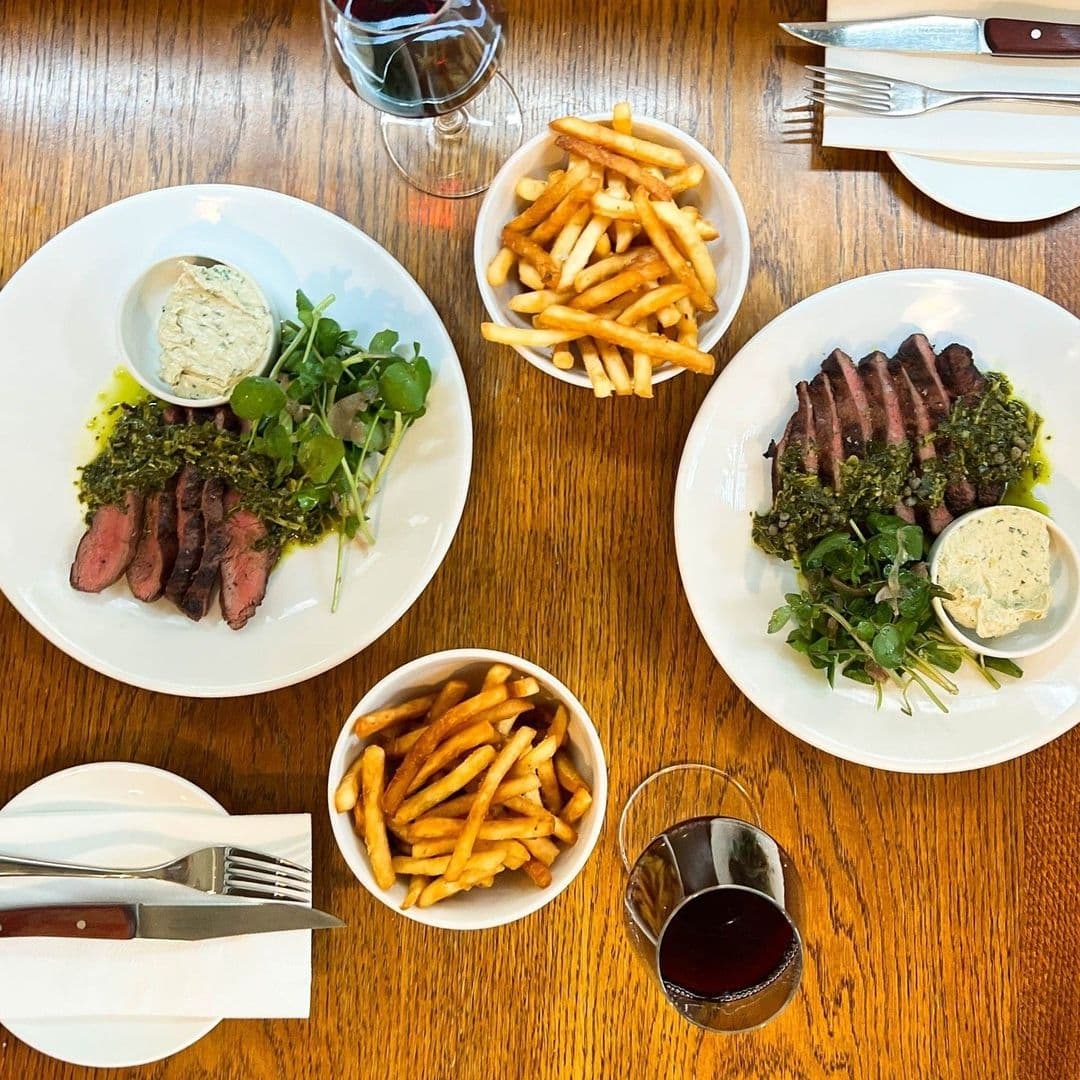 Ox Club at Headrow House Gourmet Steak Frites Dining Experience Dining experience at Ox Club at Headrow House featuring two plates of sliced steak with green salad and herb sauce, bowls of French fries, and glasses of red wine on a wooden table.