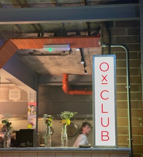 Ox Club at Headrow House Indoor Signage Cozy Ambience Flowers Illuminated vertical sign reading 'OX CLUB' at Headrow House with industrial-style interior and vases of yellow flowers on a counter.