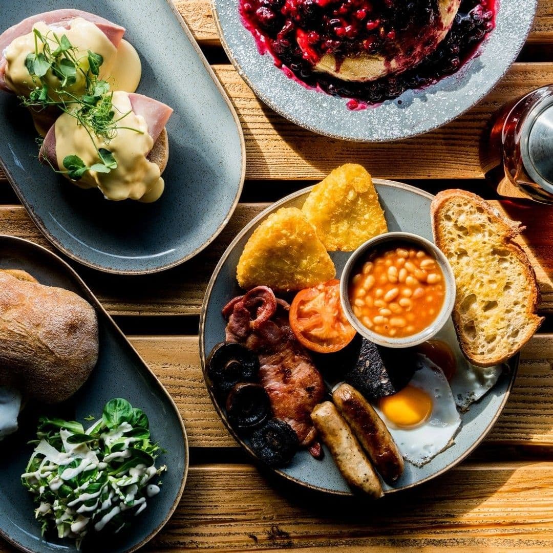 Residence Bar Cafe Kitchen Headingley Brunch Platter Sunny Table Alt text: "Delicious breakfast spread at Residence Bar Cafe | Kitchen (Headingley) featuring eggs benedict, a classic English breakfast with eggs, bacon, sausages, black pudding, hash browns, baked beans, grilled tomato, toast, and a side salad."