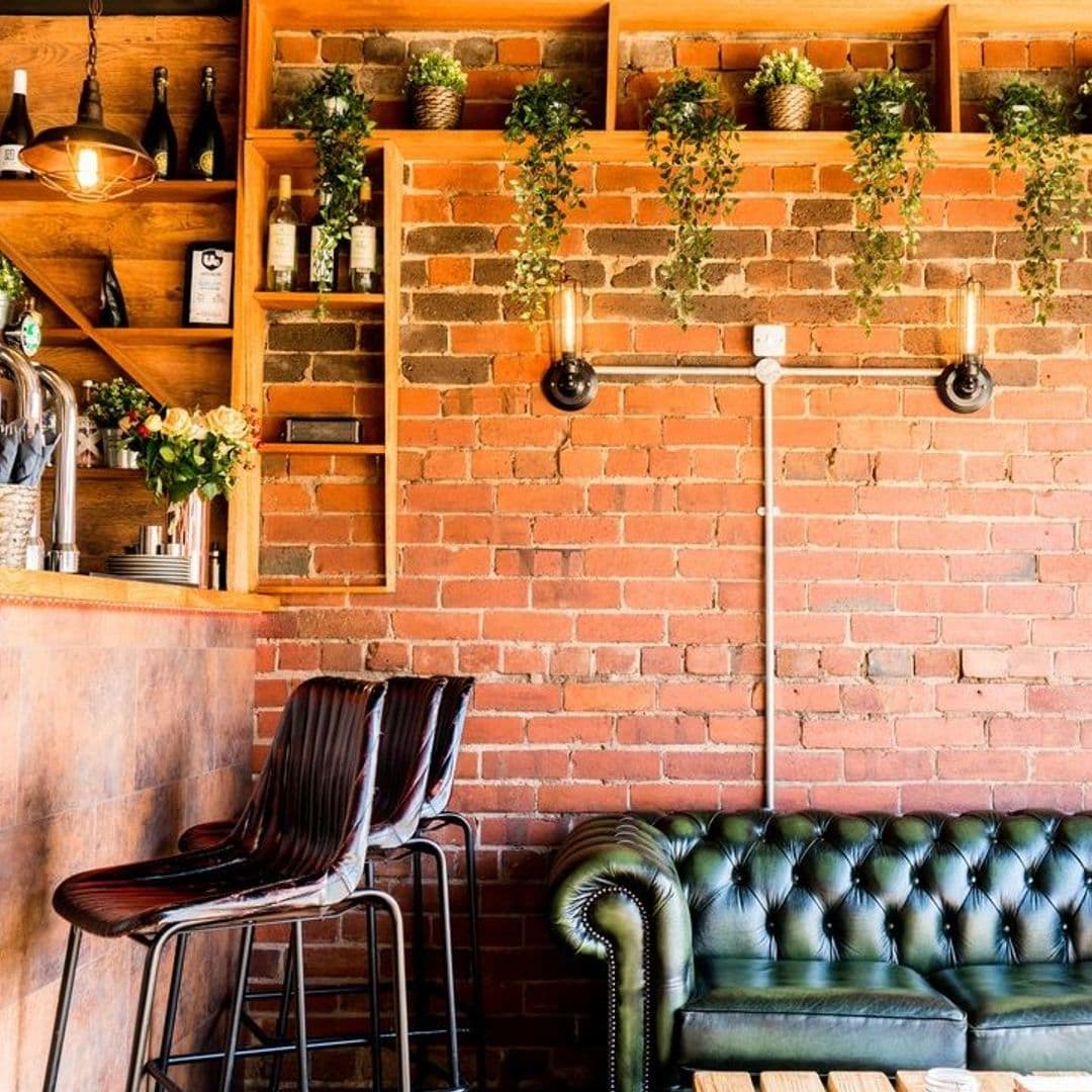 Residence Bar Cafe Kitchen Headingley Cozy Brick Interior Greenery Decor Cozy corner at Residence Bar Cafe | Kitchen (Headingley) with exposed brick walls, green leather seating, and wooden shelves with plants and bottles.