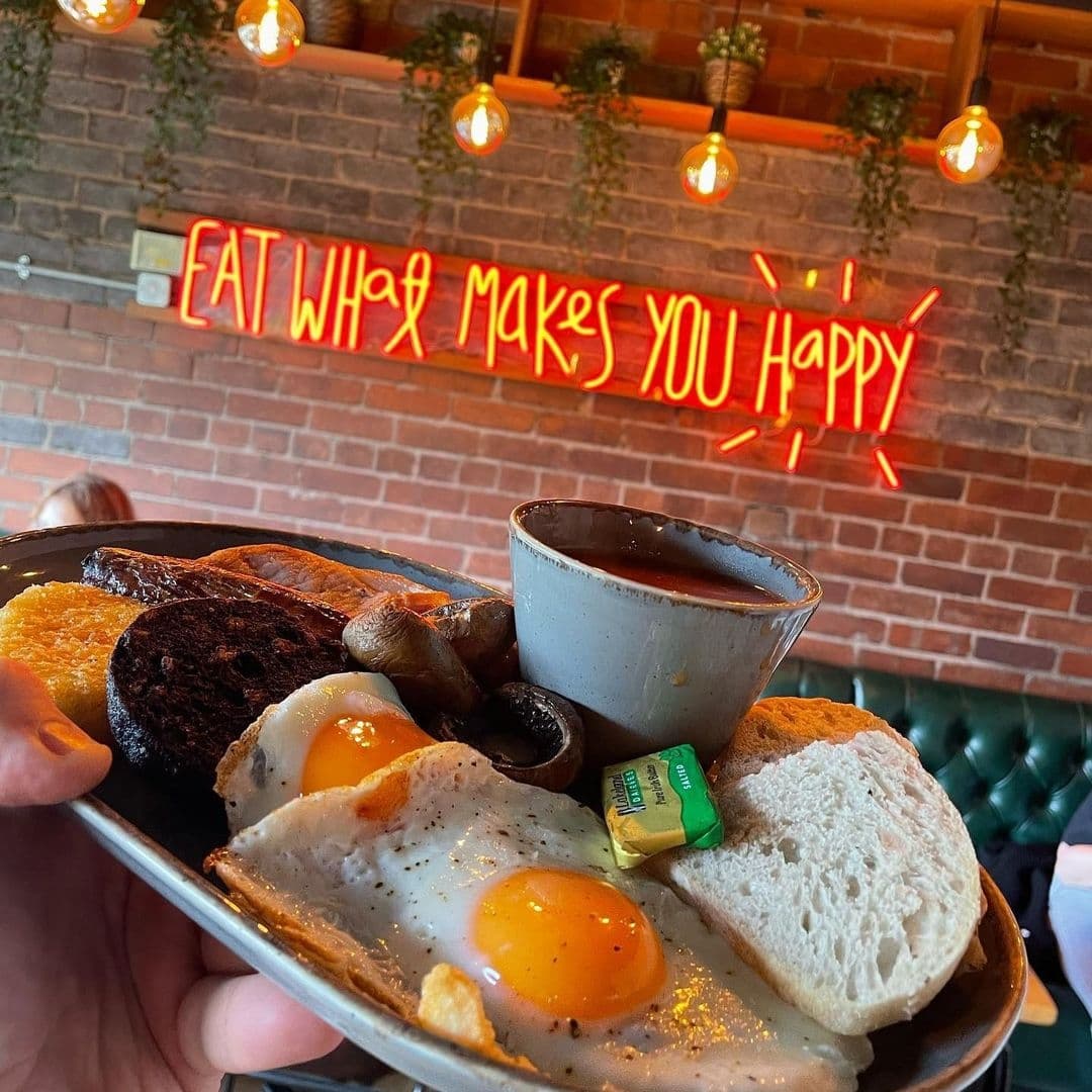 Residence Bar Cafe Kitchen Headingley Hearty Breakfast Neon Sign Brick Wall A close-up of a traditional English breakfast served on a plate, with sunny-side-up eggs, toast, black pudding, hash brown, mushrooms, and a cup of beans, at Residence Bar Cafe | Kitchen (Headingley). In the background, a neon sign reads "EAT WHAT MAKES YOU HAPPY" against a brick wall with hanging light bulbs and potted plants.