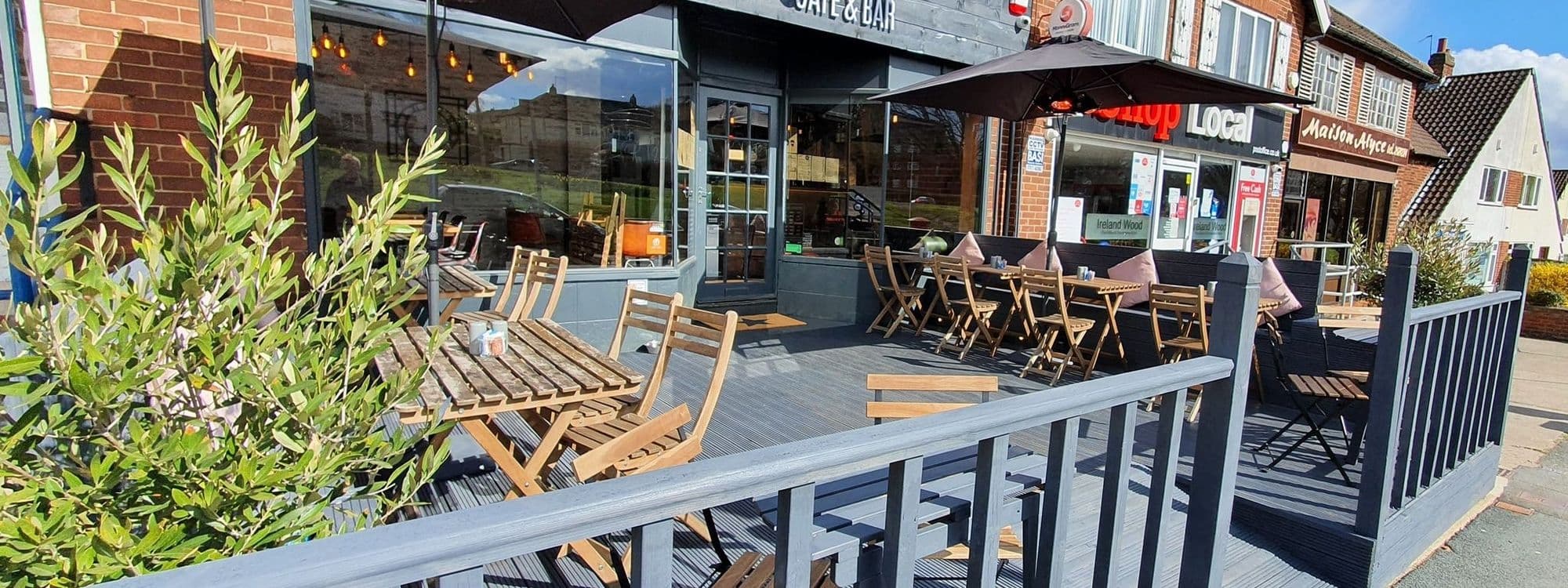 Outdoor seating area at Residence Bar Cafe | Kitchen (Headingley) with wooden tables and chairs, large umbrellas for shade, and green plants along the perimeter on a sunny day.