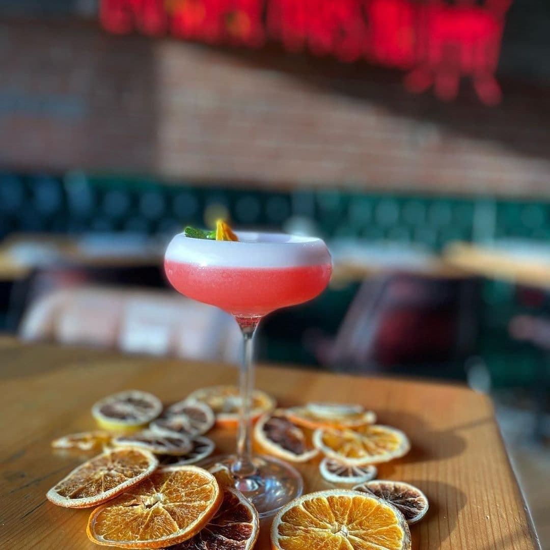 Residence Bar Cafe Kitchen Headingley_Signature Cocktail Citrus Garnish Elegant pink cocktail garnished with lime on a table at Residence Bar Cafe | Kitchen (Headingley), surrounded by dried orange slices, with cozy seating and warm lighting in the background.