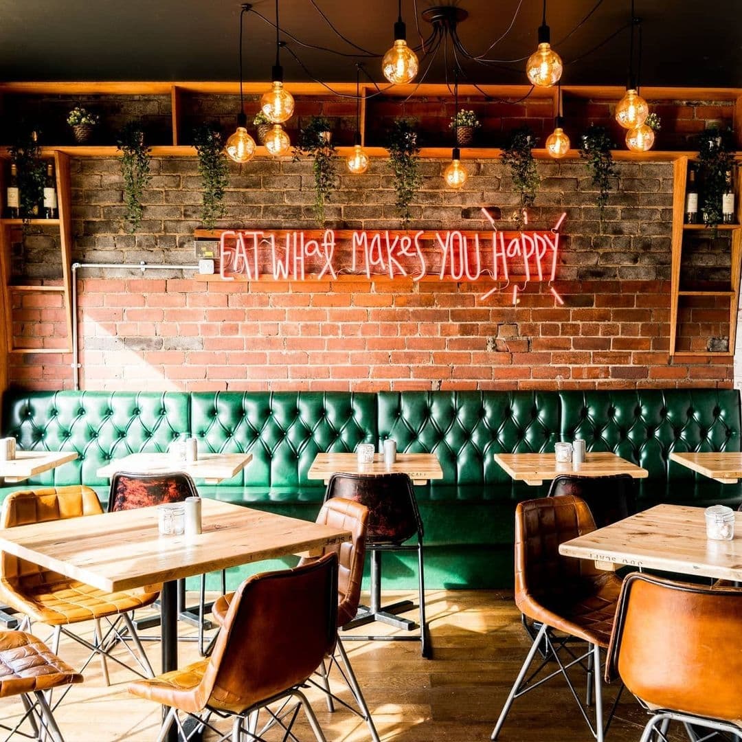 Residence_Bar_Cafe_Kitchen_Headingley_Stylish_Interior_Brick_Wall_Neon_Sign Interior of Residence Bar Cafe | Kitchen (Headingley) featuring a cozy dining area with green tufted booth seating, wooden tables, and leather chairs under warm pendant lighting. Exposed brick walls adorned with neon sign reading "EAT WHAT MAKES YOU HAPPY" and shelves with green plants create a welcoming atmosphere.