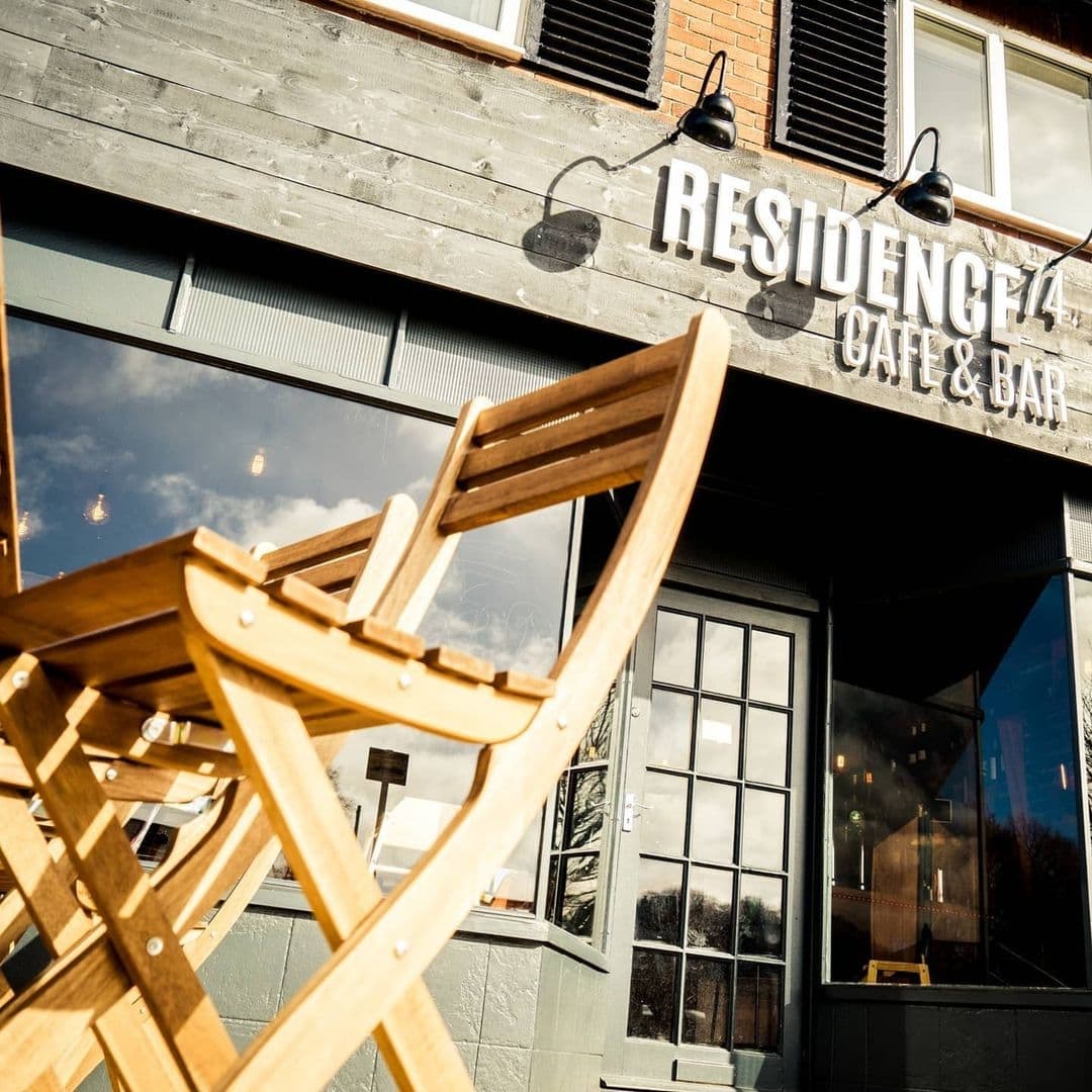 Residence_Bar_Cafe_Kitchen_Headingley_Sunny_Exterior_Wooden_Chairs Exterior view of Residence Bar Cafe | Kitchen (Headingley) with a wooden chair in the foreground and the cafe's signboard visible above the entrance.