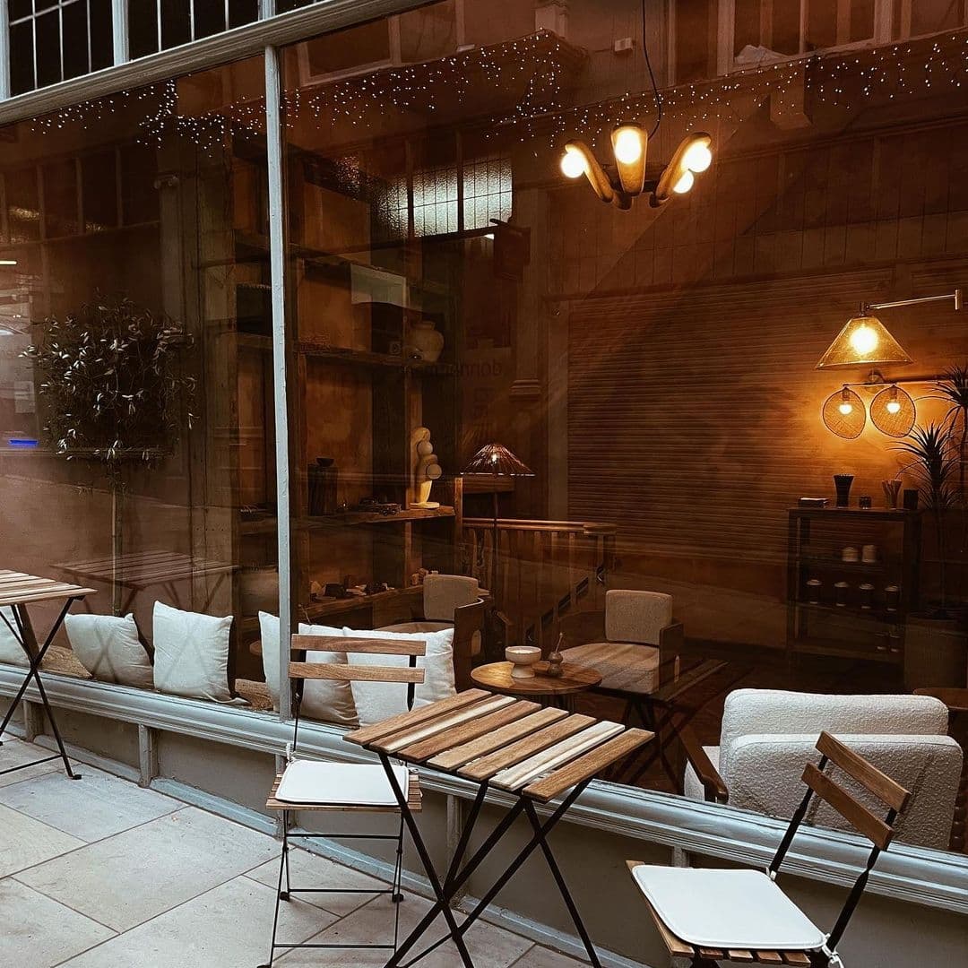 Sonder_CozyCafeEveningAmbience Cozy outdoor seating at Sonder with wooden tables, white cushioned chairs, and warm interior lighting visible through glass windows, complemented by twinkling string lights above.