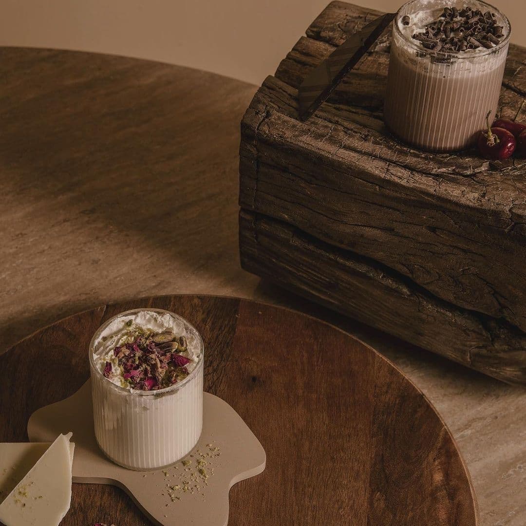 Sonder_Gourmet_Desserts_Elegant_Presentation Gourmet desserts on a wooden table at Sonder, featuring a glass of chocolate shavings topped beverage and another with a floral garnish, with a rustic wooden backdrop.