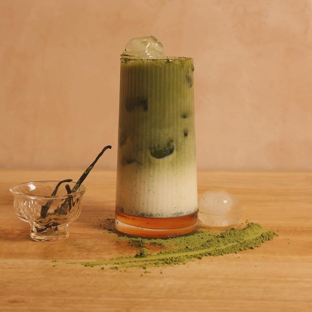 Sonder_Matcha_Latte_Artisan_Drink A tall glass of layered matcha drink with ice on top, a sprinkle of matcha powder on the wooden surface, and a vanilla pod in a glass dish at Sonder.