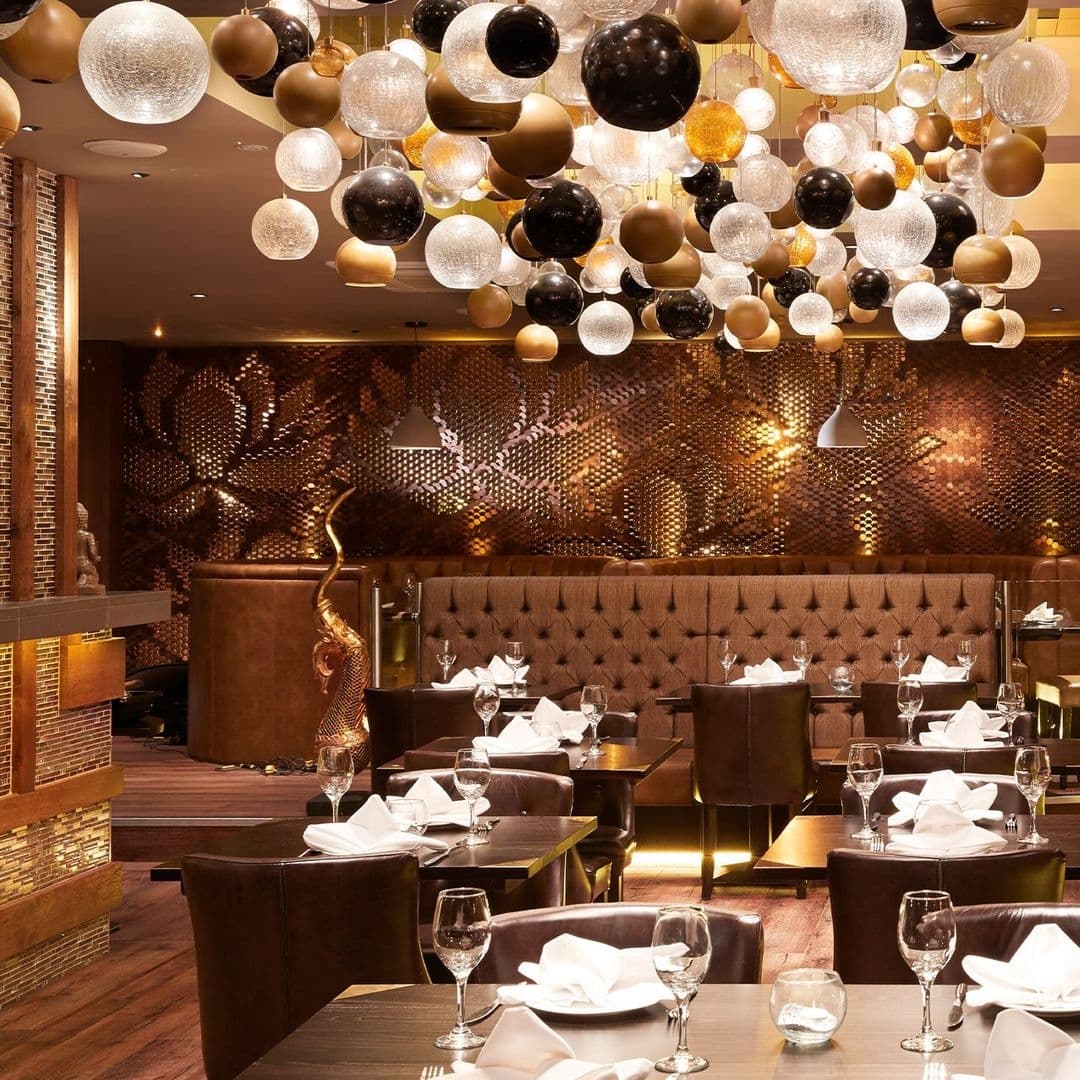 Sukhothai Chapel Allerton Elegant Dining Interior Decor Elegant interior of Sukhothai Chapel Allerton restaurant with a ceiling adorned with an array of spherical and globe pendant lights in various sizes and colors, over neatly arranged dining tables with white napkins and wine glasses. The walls feature intricate, patterned designs adding to the luxurious ambiance.