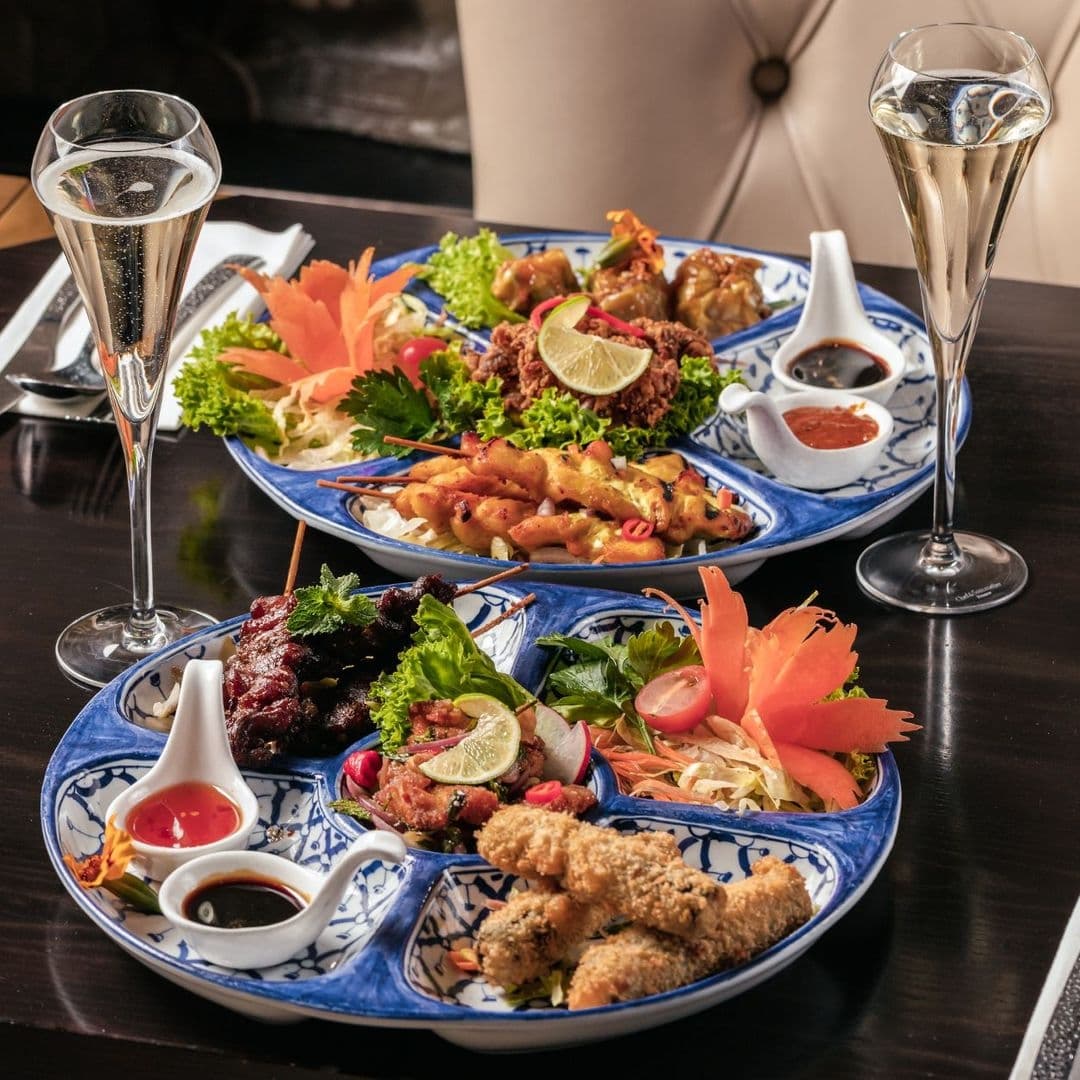 Sukhothai Chapel Allerton Thai Cuisine Dining Experience Assorted Thai dishes on blue patterned plates at Sukhothai Chapel Allerton, including skewered meats, fried appetizers, and fresh salads, accompanied by sauces and two glasses of sparkling wine.