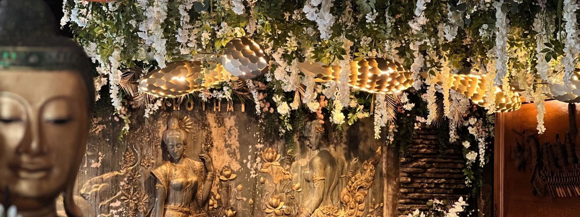 Interior decor of Sukhothai Chapel Allerton featuring golden Buddha statues, intricate wall carvings, and hanging floral arrangements with warm ambient lighting.