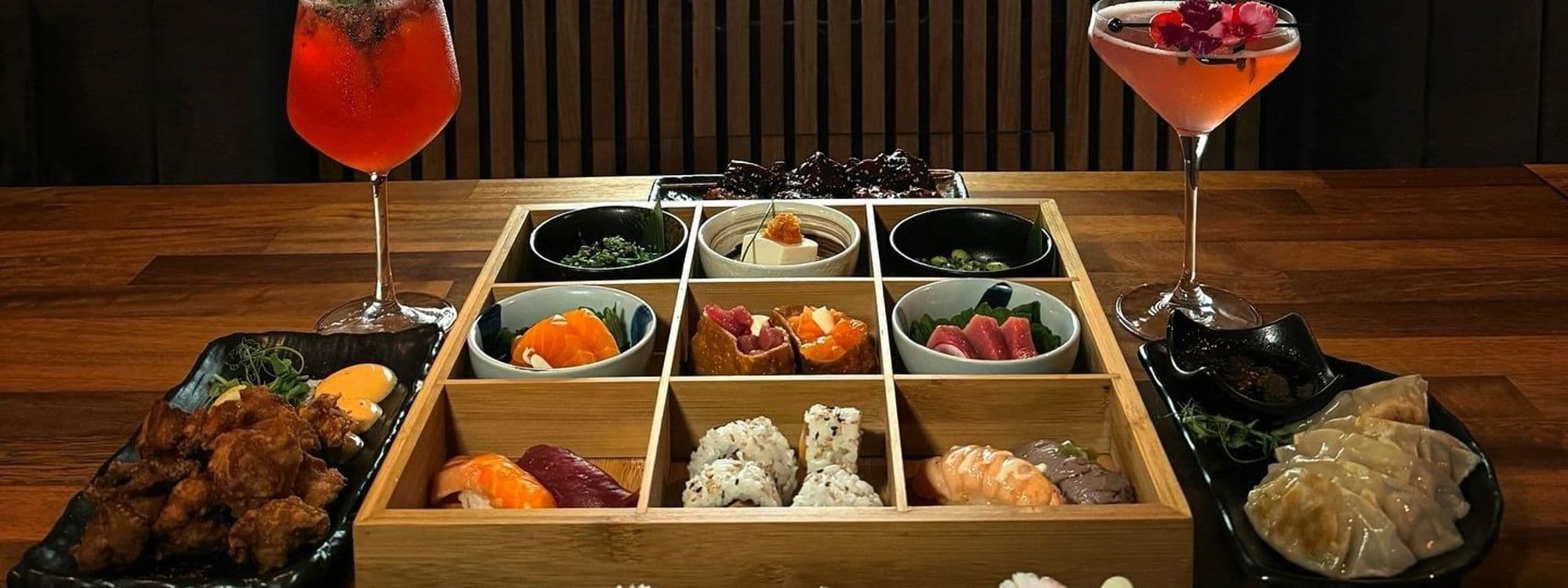Assorted Japanese cuisine at TADA featuring sushi, sashimi, dumplings, and fried chicken, complemented by colorful cocktails, presented on a wooden table with a bamboo backdrop.