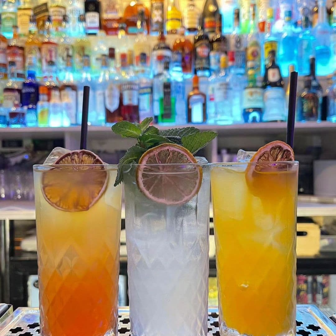 Tavassolis Cafe and Grill Colorful Cocktails Bar Backdrop Three colorful cocktails on the bar at Tavassoli's Cafe and Grill, with a backdrop of various bottles of alcohol.