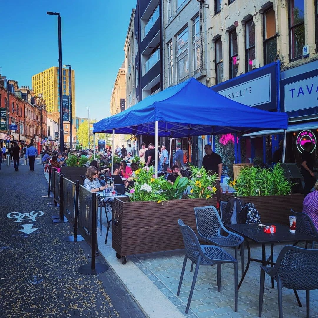 Tavassolis Cafe and Grill Outdoor Dining Bustling Street Blue Canopy Outdoor seating at Tavassoli's Cafe and Grill with patrons dining under a blue canopy, surrounded by potted plants, on a sunny day in a bustling city street.