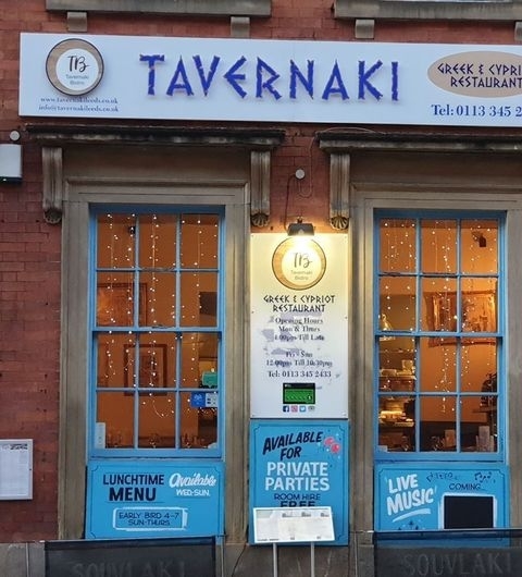 Tavernaki Bistro Evening Ambience Greek Cypriot Restaurant Exterior view of Tavernaki Bistro, showcasing its warm, inviting facade with blue framed windows, glowing interior lights, and signage promoting Greek and Cypriot cuisine, available for private parties, and live music events.
