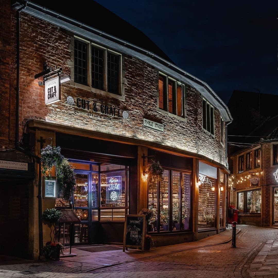 The Cut and Craft Evening Ambiance Steakhouse Brick Building Exterior night view of The Cut & Craft, a cozy steakhouse with illuminated signage and warm window lighting, set in a traditional brick building on a cobblestone street.