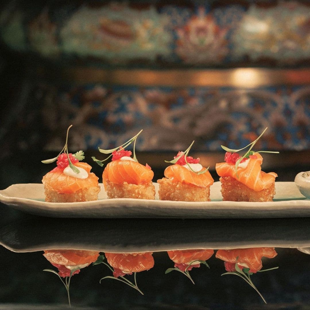 The Ivy Asia_Elegant Sushi Platter Gourmet Reflection Elegant sushi selection served at The Ivy Asia, featuring fresh salmon on crispy rice with delicate garnishes, presented on a ceramic plate with a reflective surface creating a symmetrical image.