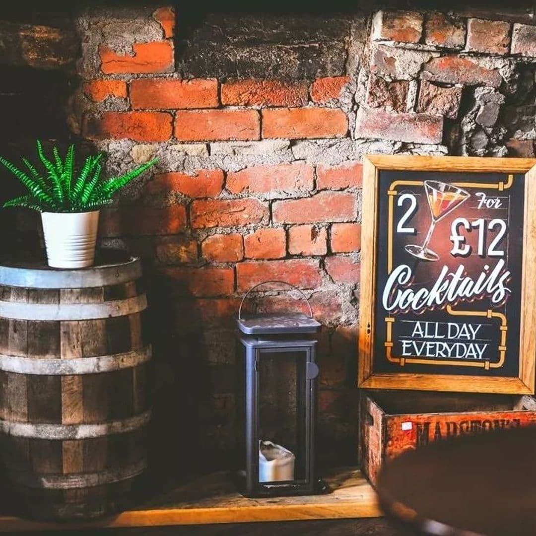 The Malt Brewhouse Cocktail Specials Brick Wall Barrel Decor Rustic corner at The Malt Brewhouse featuring a wooden barrel, a chalkboard sign with cocktail offers, and a potted green plant against an exposed brick wall.
