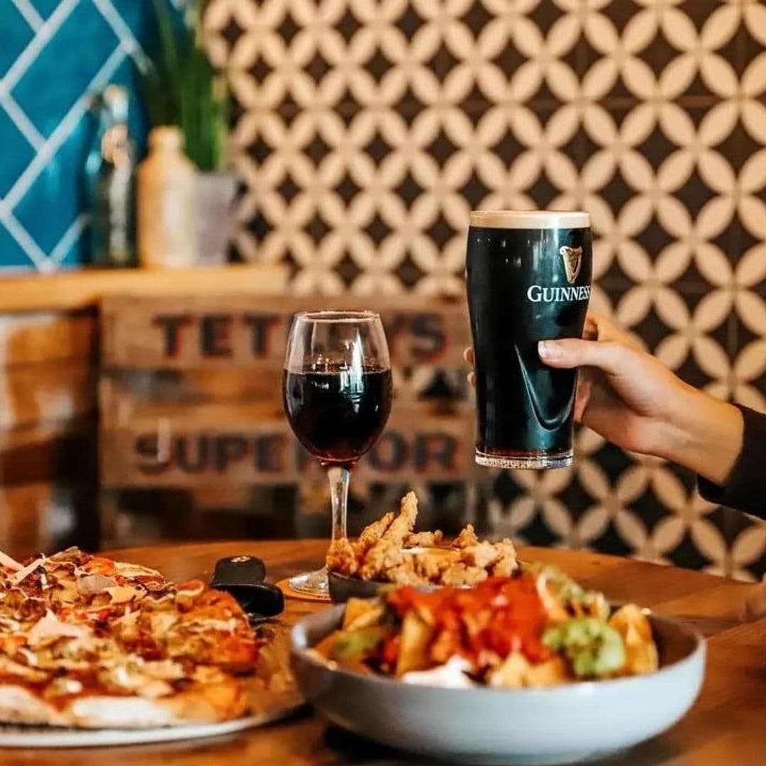 The Malt Brewhouse Craft Beer Pizza Dining Experience A person holding a pint of Guinness with a glass of red wine, a pizza, and a plate of appetizers on a table at The Malt Brewhouse.