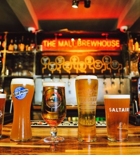 The Malt Brewhouse Craft Beer Taps Pints Bar Atmosphere Alt text: "A variety of craft beers on the bar counter at The Malt Brewhouse, with the illuminated sign of the establishment in the background."