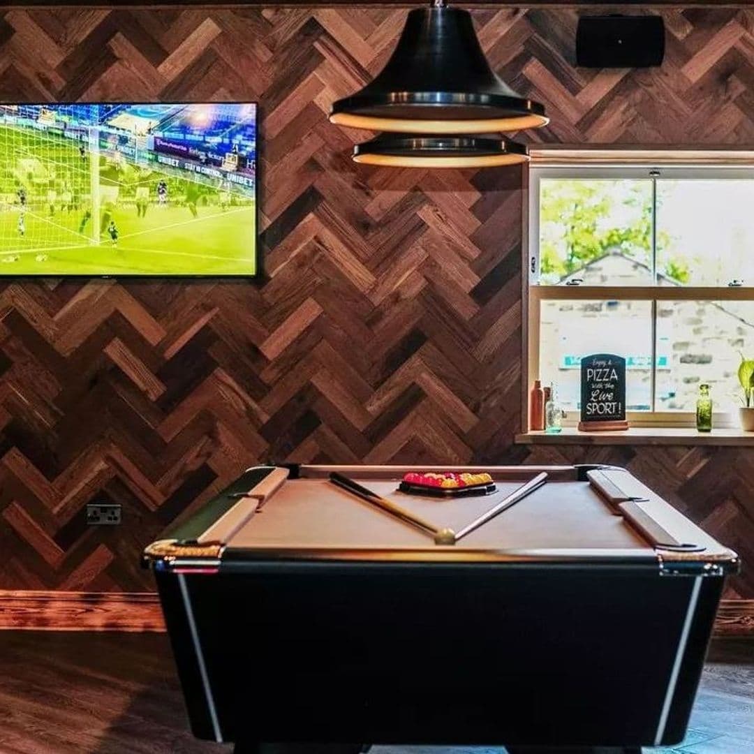 The Malt Brewhouse Pool Table Sports TV Relaxing Ambiance Interior of The Malt Brewhouse featuring a pool table with cues and balls, herringbone-patterned wood wall, pendant light, and a TV screen showing sports. A window with a "Pizza Love Sport" sign is visible in the background.