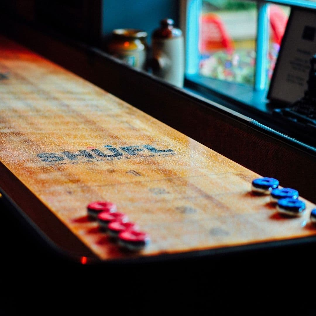The Malt Brewhouse_Vintage Shuffleboard Game Warm Ambiance Close-up of a shuffleboard table with blue and red pucks at The Malt Brewhouse, with a warm ambiance and a window in the background showing outdoor greenery.