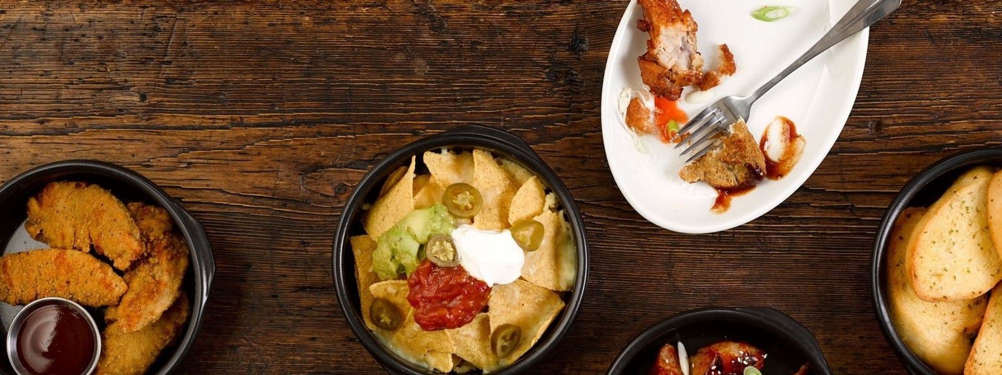 Assorted appetizers at The Merchant featuring crispy chicken tenders with dipping sauce, nachos with jalapenos, guacamole, sour cream, and salsa, and garlic bread on a rustic wooden table.
