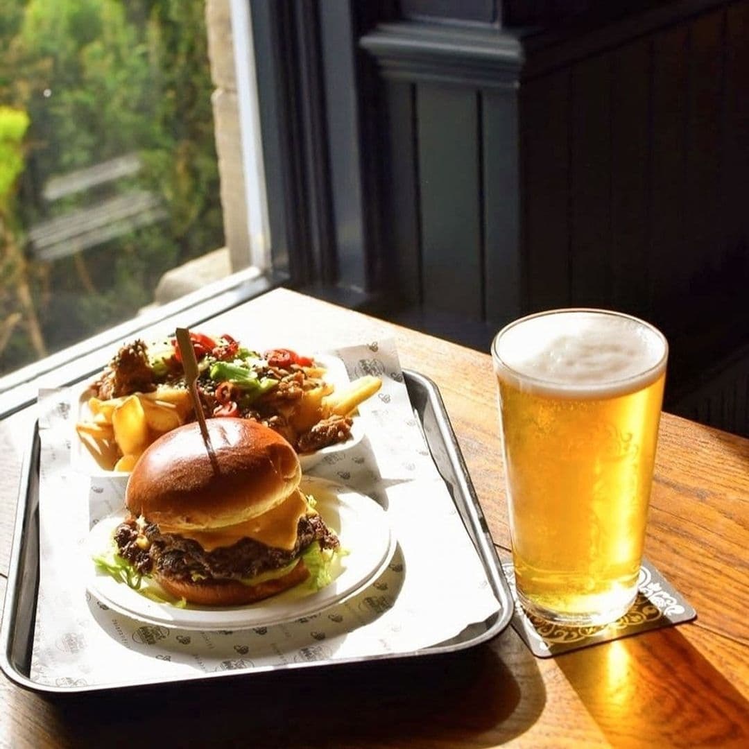 The Old Kings Arms_Gourmet_Burger_Cold_Beer_Window_View Appetizing cheeseburger with toppings and fries on a tray next to a pint of beer, with natural light illuminating the meal at The Old Kings Arms.