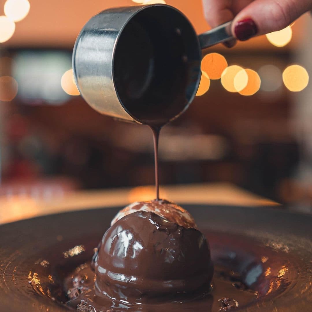 The Whitehall Restaurant Bar Chocolate Dessert Pouring Sauce Bokeh Lights Decadent chocolate dessert being served at The Whitehall Restaurant & Bar, with warm chocolate sauce poured over a spherical chocolate cake on a plate, against a softly lit background.