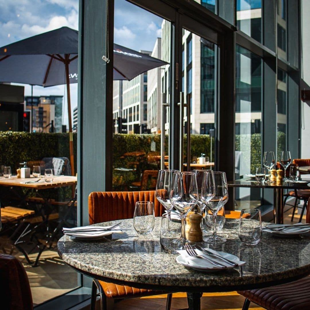 The Whitehall Restaurant Bar Elegant Outdoor Dining Setting Sunny Day Outdoor dining setup at The Whitehall Restaurant & Bar with a marble table set with wine glasses, cutlery, and a pepper mill, overlooking an urban landscape with modern buildings and a blue sky.