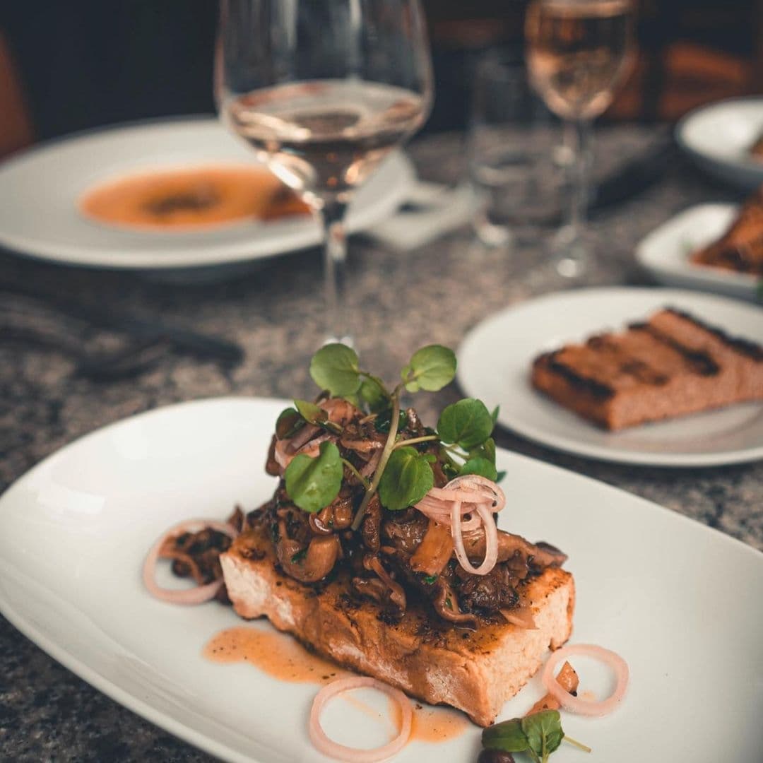 The Whitehall Restaurant Bar Gourmet Entree Wine Dining Experience Gourmet dish featuring grilled salmon topped with mushrooms and fresh greens, garnished with onion rings, served at The Whitehall Restaurant & Bar, with a glass of rosé wine in the background.