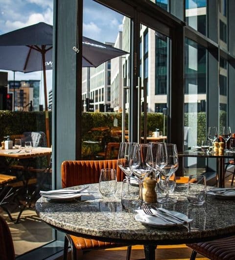 The Whitehall Restaurant Bar Upscale Dining Patio View Elegant dining setup at The Whitehall Restaurant & Bar with wine glasses on a marble table, plush seating, and a view of the cityscape.