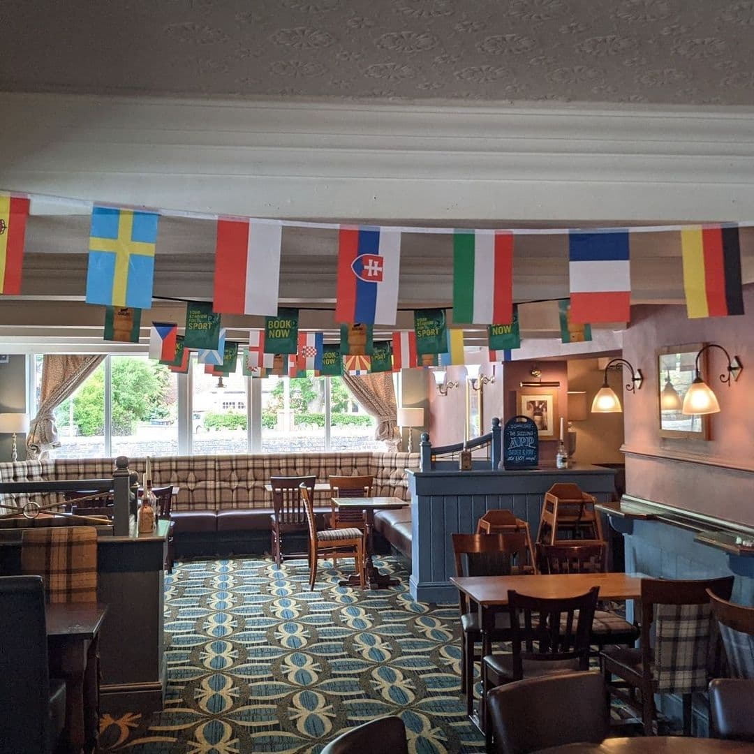 The Woodcock New Farnley International Flags Pub Interior Decor Interior of The Woodcock New Farnley pub with national flags hanging across the ceiling, traditional wooden furniture, patterned carpet, and a well-lit bar area.