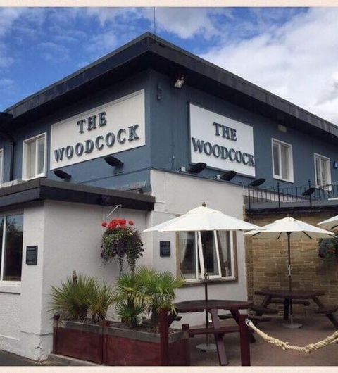 The_Woodcock_New_Farnley_Pub_Exterior_Sunny_Day Exterior view of The Woodcock New Farnley, featuring the building's blue and white facade with signage, outdoor seating with parasols, and hanging flower baskets.