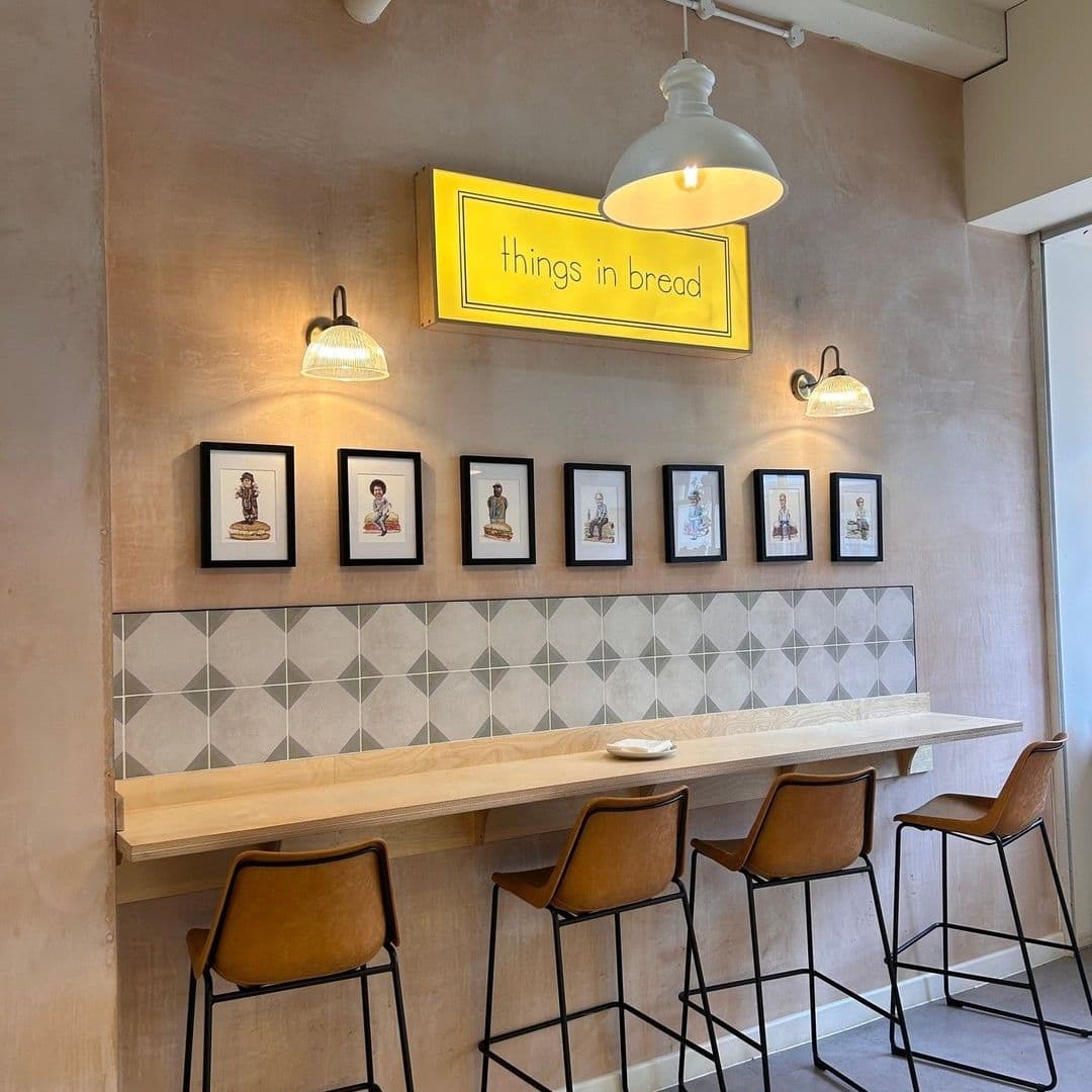 Things in Bread cafe interior design modern wooden furniture Interior of "Things in Bread" featuring a minimalist dining area with a long wooden counter, brown leather stools, geometric-patterned tile backsplash, and framed artwork on the wall, illuminated by pendant lights and a bright yellow sign with the establishment's name.