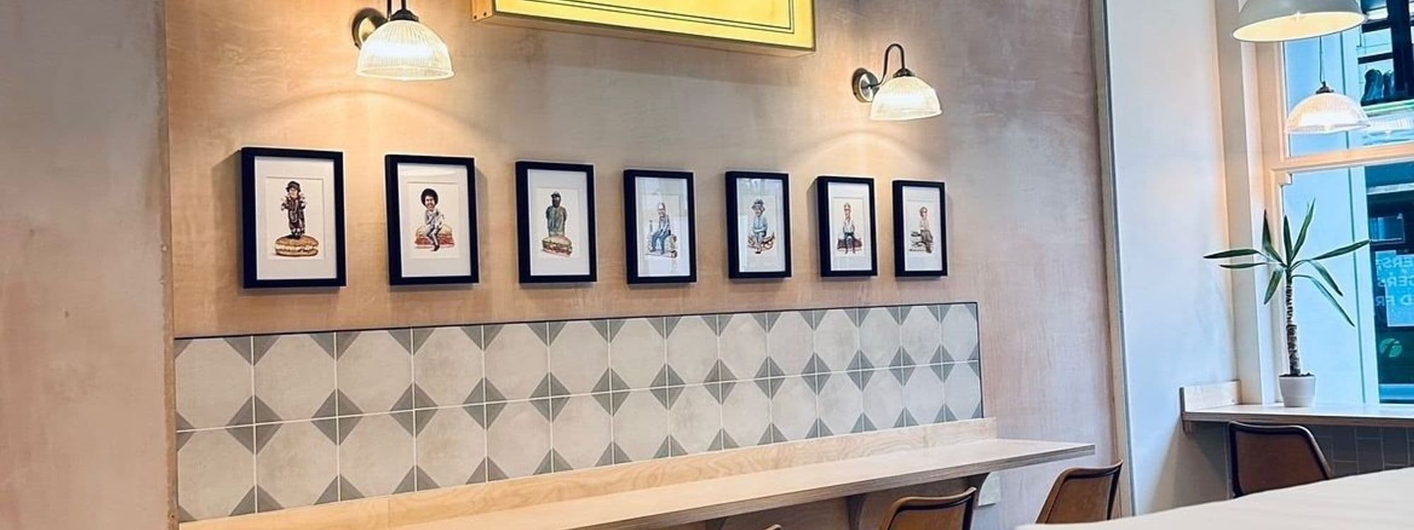 Interior of Things in Bread featuring a row of framed illustrations above a bench with geometric tile backsplash, pendant lighting, and a potted plant.