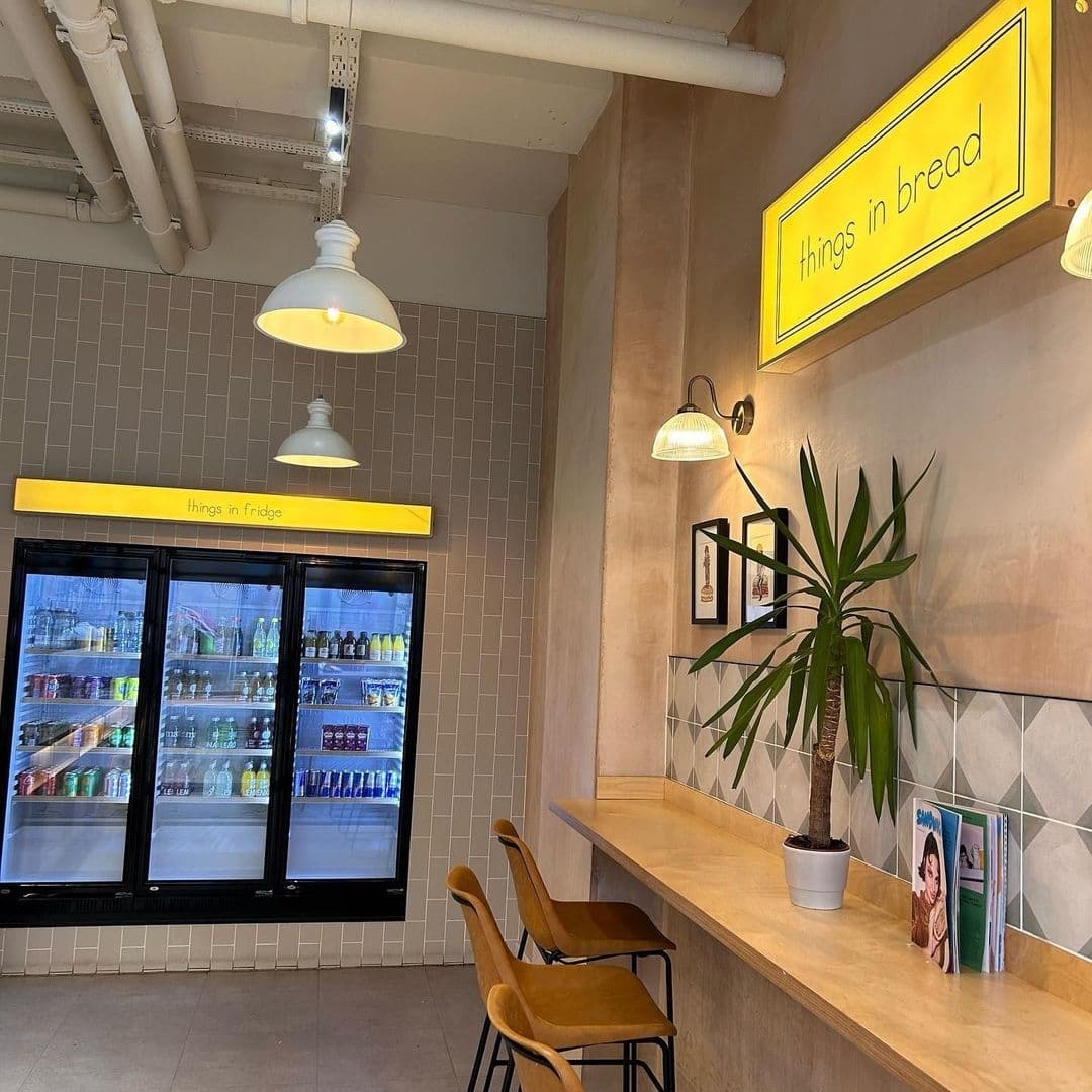 Things_in_Bread_Cafe_Interior_Design_Beverage_Fridge Interior view of Things in Bread café with a beverage fridge, wooden seating, potted plant, and wall art under warm lighting.