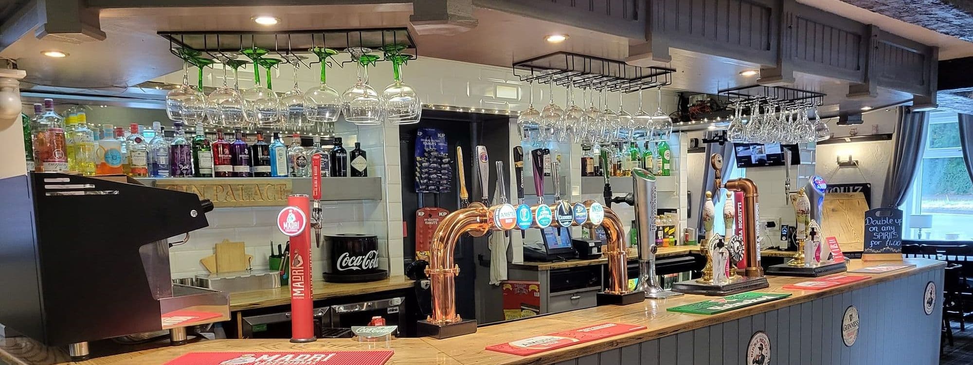 Interior view of the bar at Three Horse Shoes Oulton, showcasing a selection of draft beer taps, a variety of spirits on shelves, and wine glasses hanging from racks.