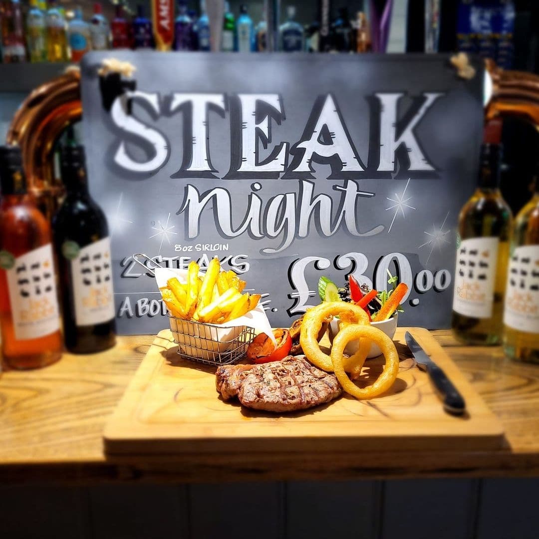 Three_Horse_Shoes_Oulton_Steak_Night_Dining_Experience Grilled steak served with fries and onion rings on a wooden board at Three Horse Shoes Oulton's Steak Night, with a promotional sign and bottles of sauce in the background.
