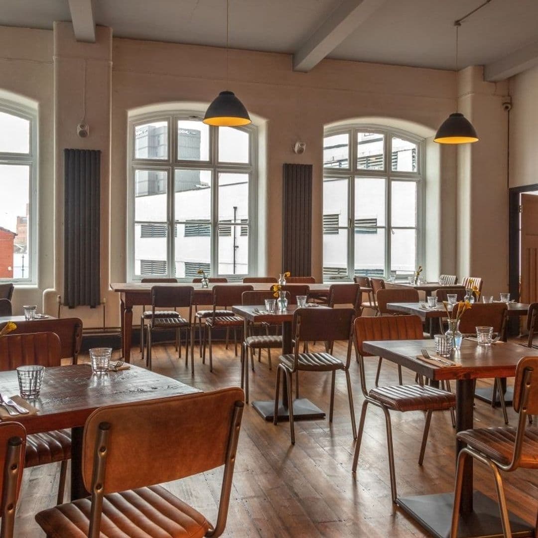 Threes A Crowd_elegant dining space natural light wooden furniture Interior of Three's A Crowd showing a spacious dining area with wooden floors, large windows, and pendant lighting. Wooden tables set with glasses and yellow flowers create a welcoming atmosphere.