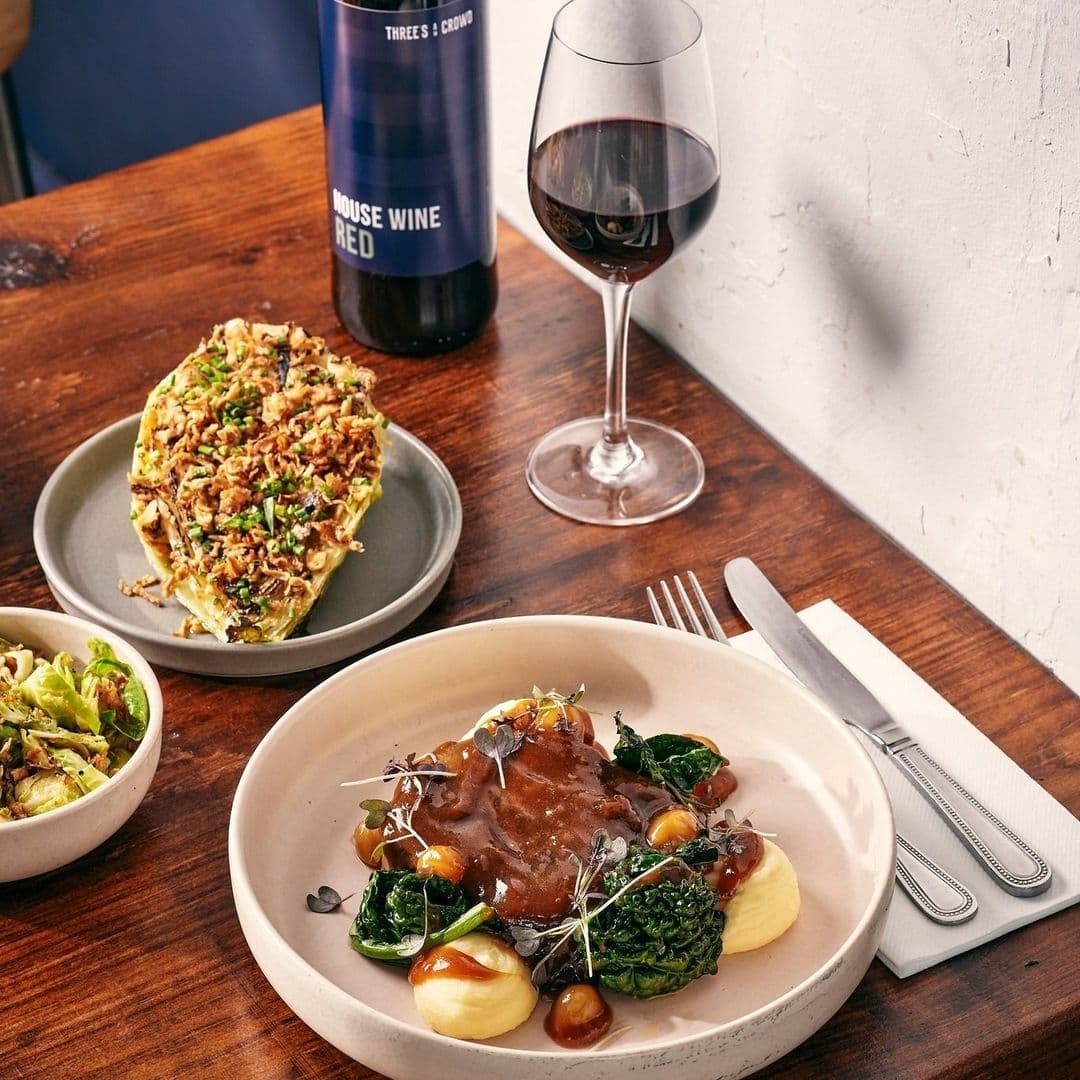 Threes A Crowd_gourmet dining wine elegant presentation Gourmet dining experience at Three's A Crowd, featuring a plate of braised meat with gravy on a bed of mashed potatoes and greens, a side of salad, a slice of savory pie, a glass of red wine, and a bottle labeled 'House Wine Red'.