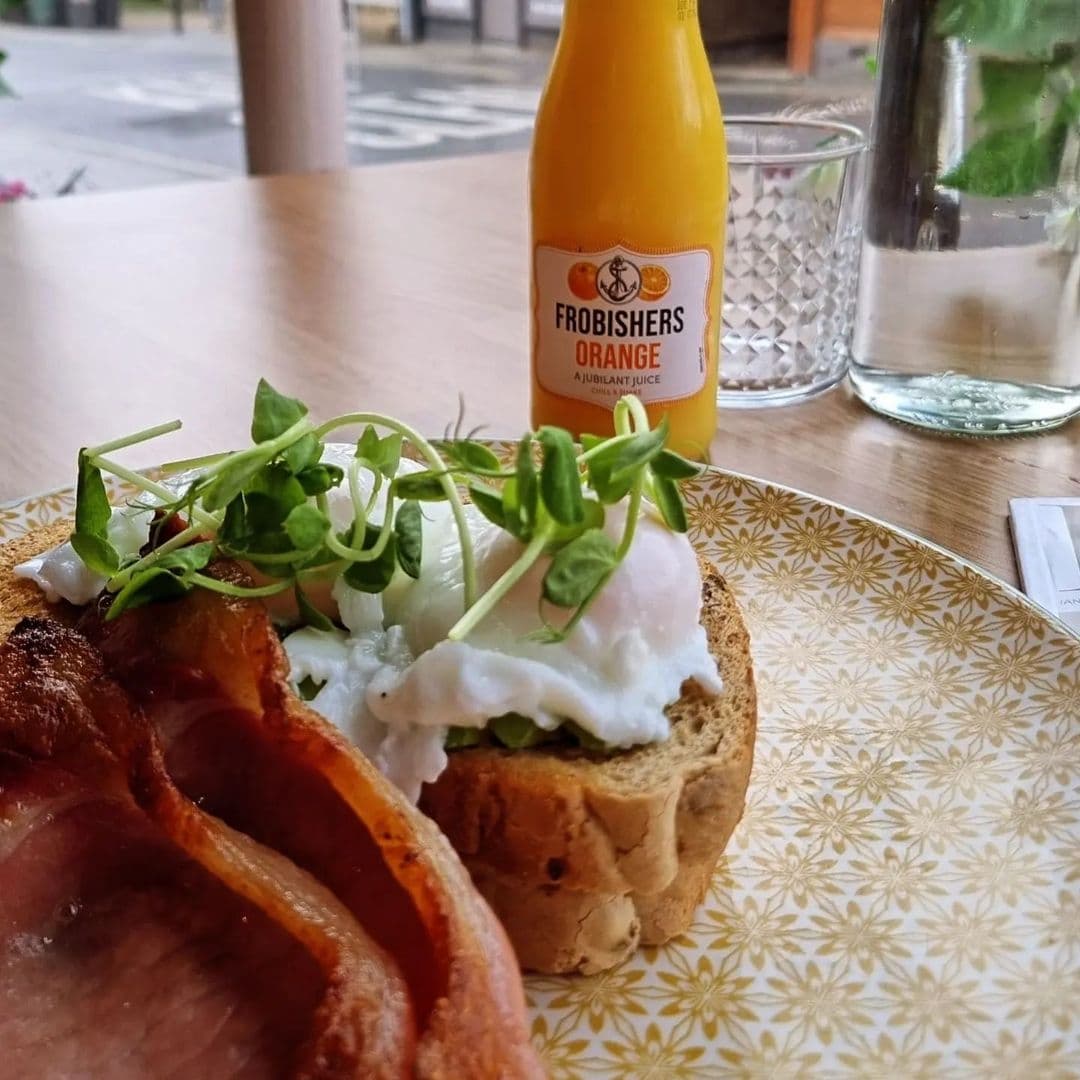 Truly Scrummy Delicious Breakfast Plate Orange Juice Alt text: "Delicious breakfast at Truly Scrummy featuring poached egg on toast with bacon, garnished with fresh greens, accompanied by a bottle of Frobishers orange juice and a glass of water with mint."