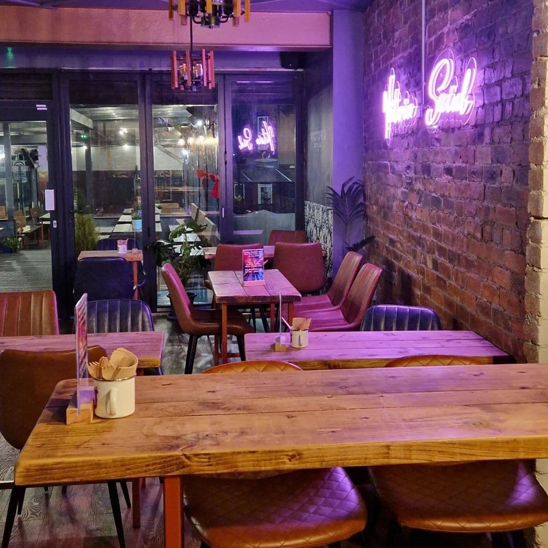 Victoria Social Neon Sign Cozy Interior Wooden Tables Interior of Victoria Social with wooden tables, purple upholstered seating, and neon signage on brick wall.